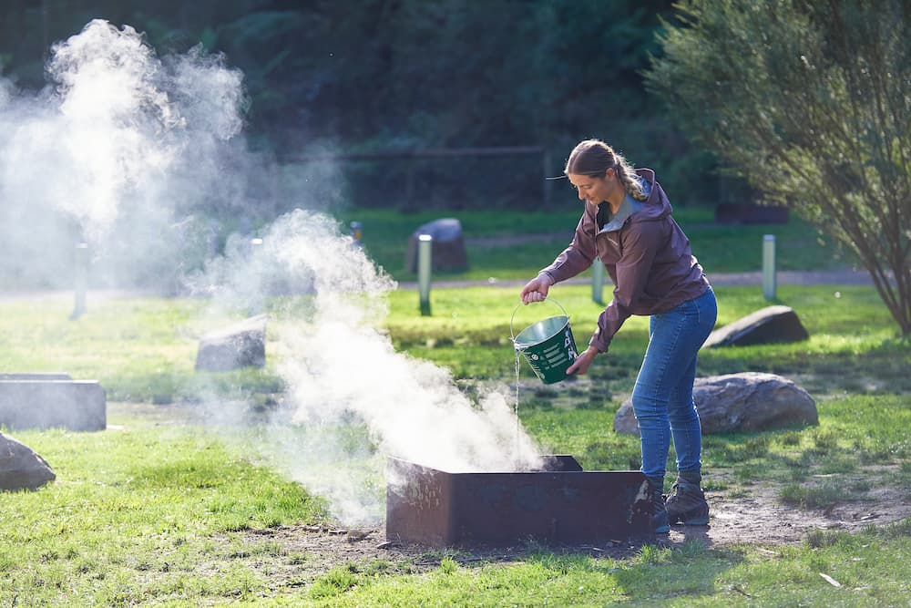 Woman in jeans and coat pouring water over a campfire