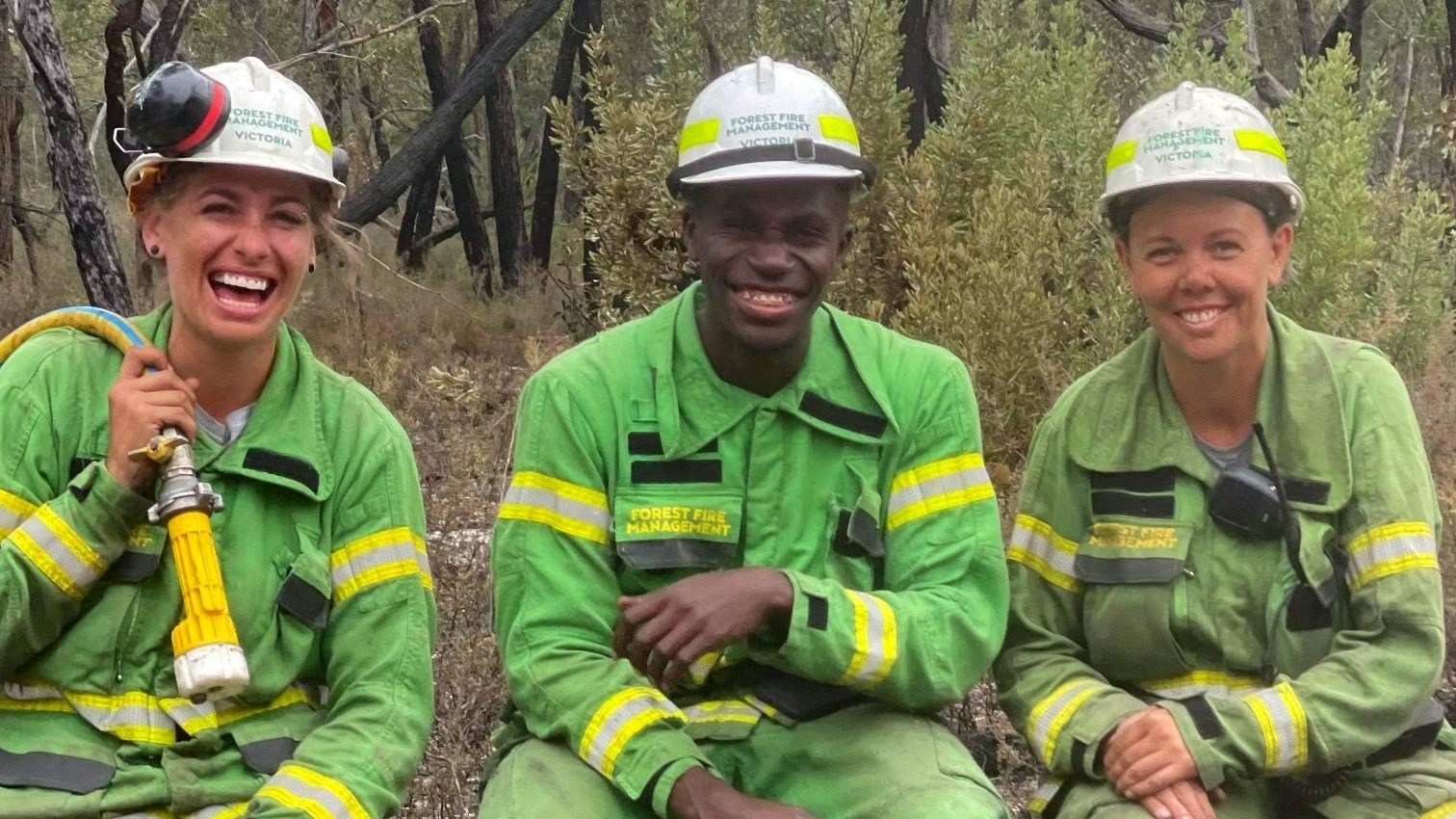 Three people in green firefighting uniforms sit in a row smiling. 