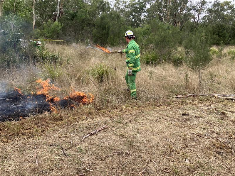 Photo of person in helmet and FFMVic greens holding drip torch with a patch of burning ground visible in front of them