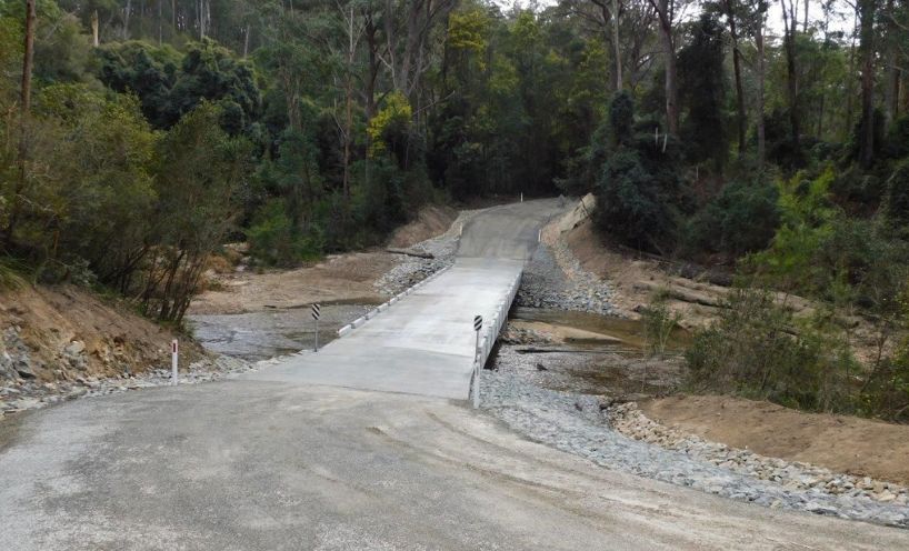 Rocky River Road bridge. Image is shot in forested country and shows a gravel road approach sloping down to the low, one-lane concrete bridge over a shallow river and a gravel road leading up from the crossing on the other side of the river.