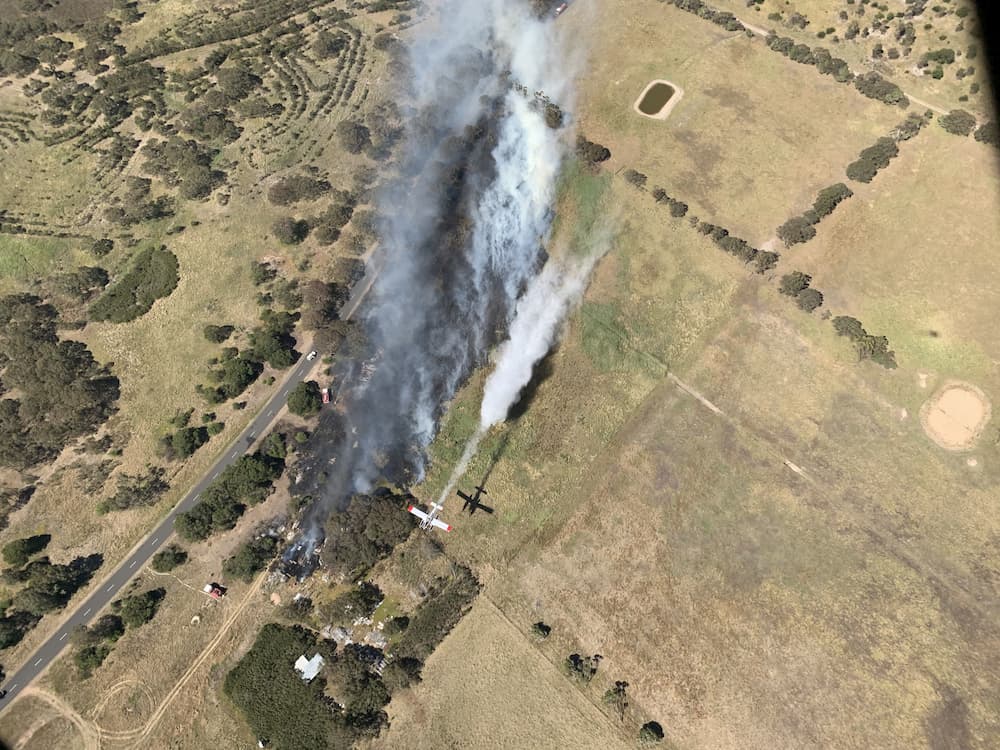Bird's eye view of fixed-wing aircraft flying over a small fire in grassland near a road. Grey smoke is billowing from the ground