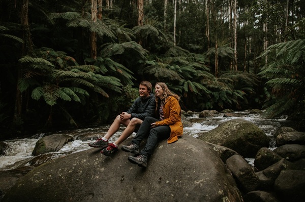 Photo of two people sitting on a large rock next to a river, with tall trees and ferns visible across the other side of the river
