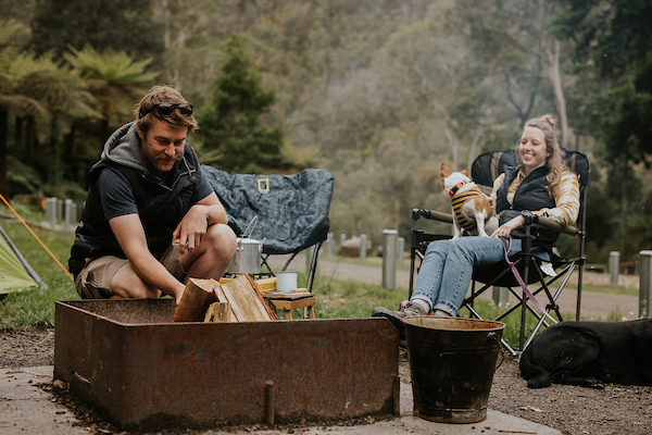 Man kneeling putting wood on a campfire, with woman sitting on camping chair, holding dog
