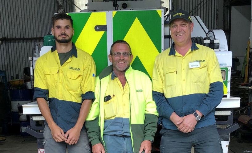 Chris Mongta (left), Chris Leary and Andy Scott, dressed in green and fluorescent yellow Forest Fire Management Victoria uniforms stand side by side in front of a slip-on ute in a corrugated iron shed at the DELWP Cann River depot.