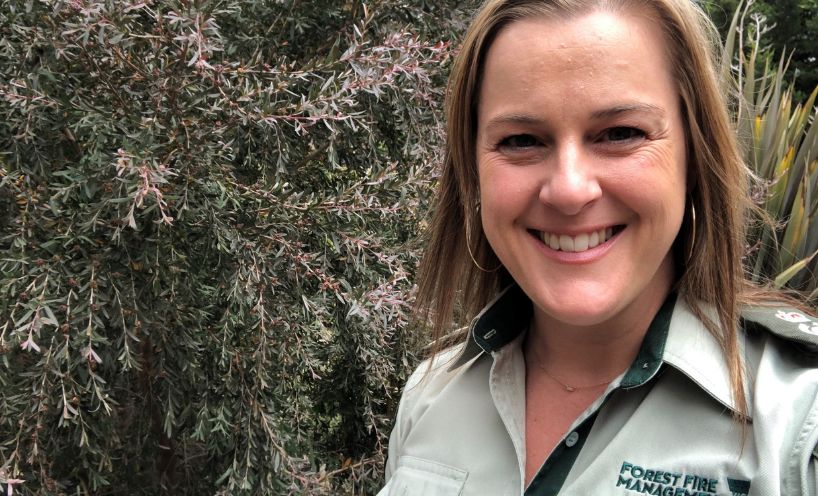 Allyson Larder, dressed in her khaki Forest Fire Management Victoria uniform shirt, smiles. She is standing to the right of the image in front of shrubs in a garden.